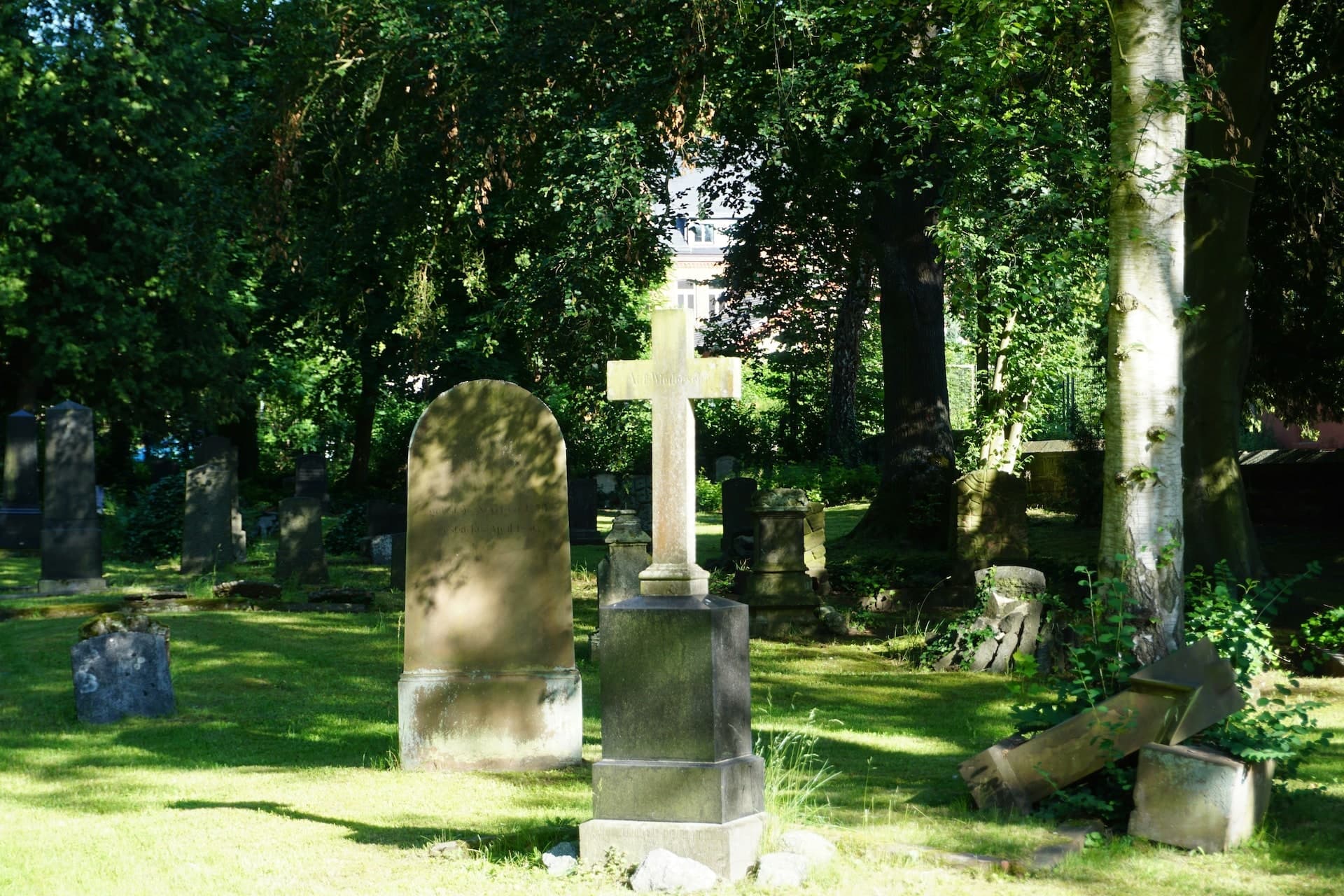 Historisches Steinkreuz auf einem schattigen Friedhof mit alten Grabsteinen und dichtem Baumbestand.