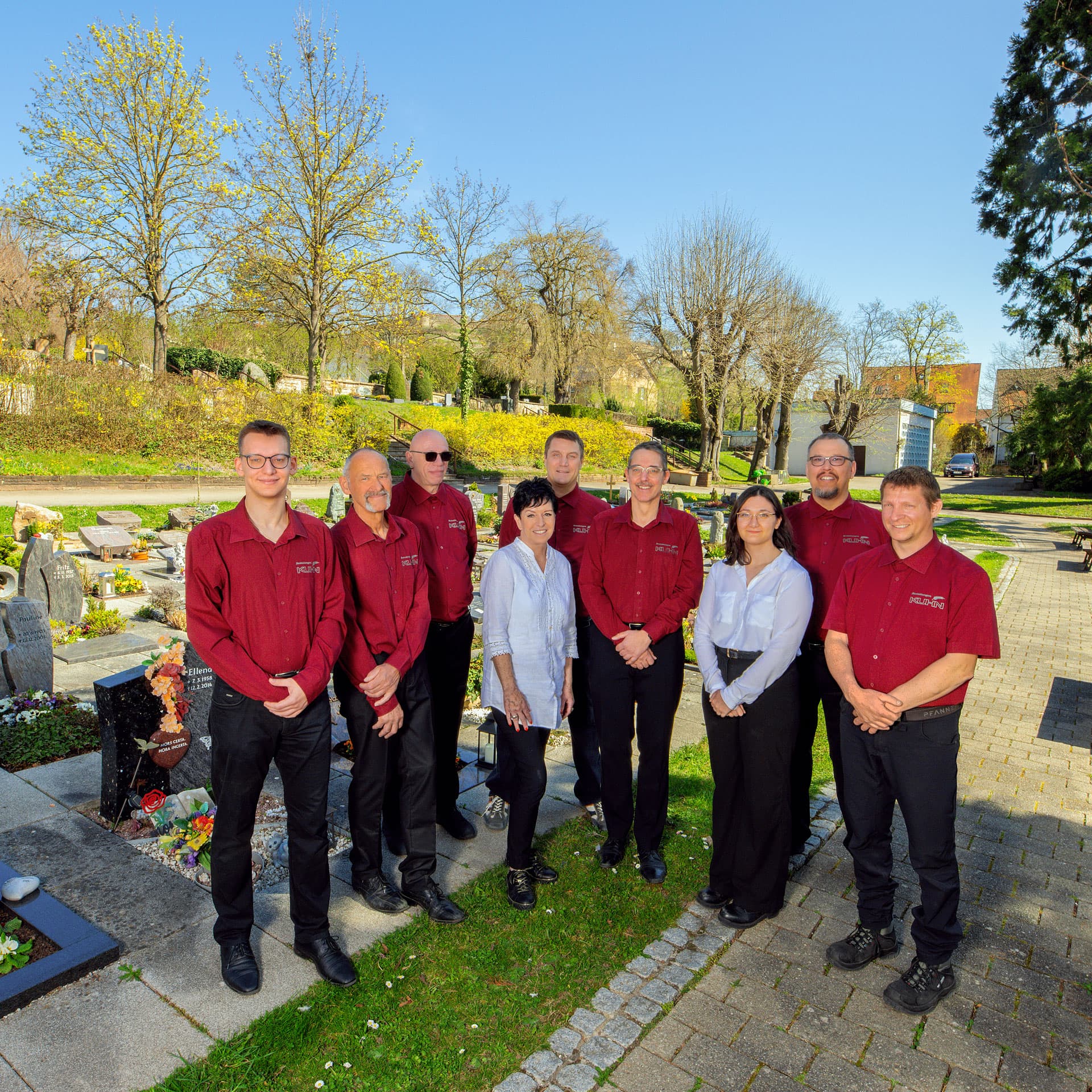 Neunköpfiges Team von Kuhn Bestattungen in dunkelroter Arbeitskleidung auf einem Friedhof bei Sonnenschein